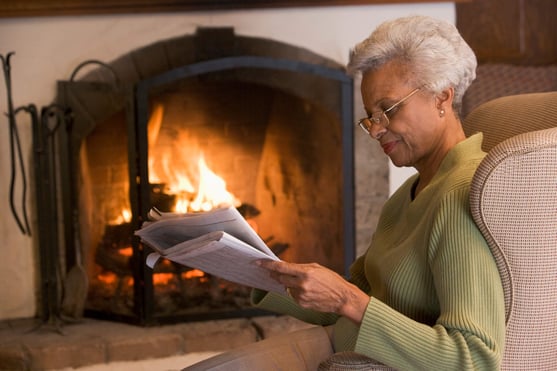 Senior woman reading a newspaper by the fire to keep warm in the winter