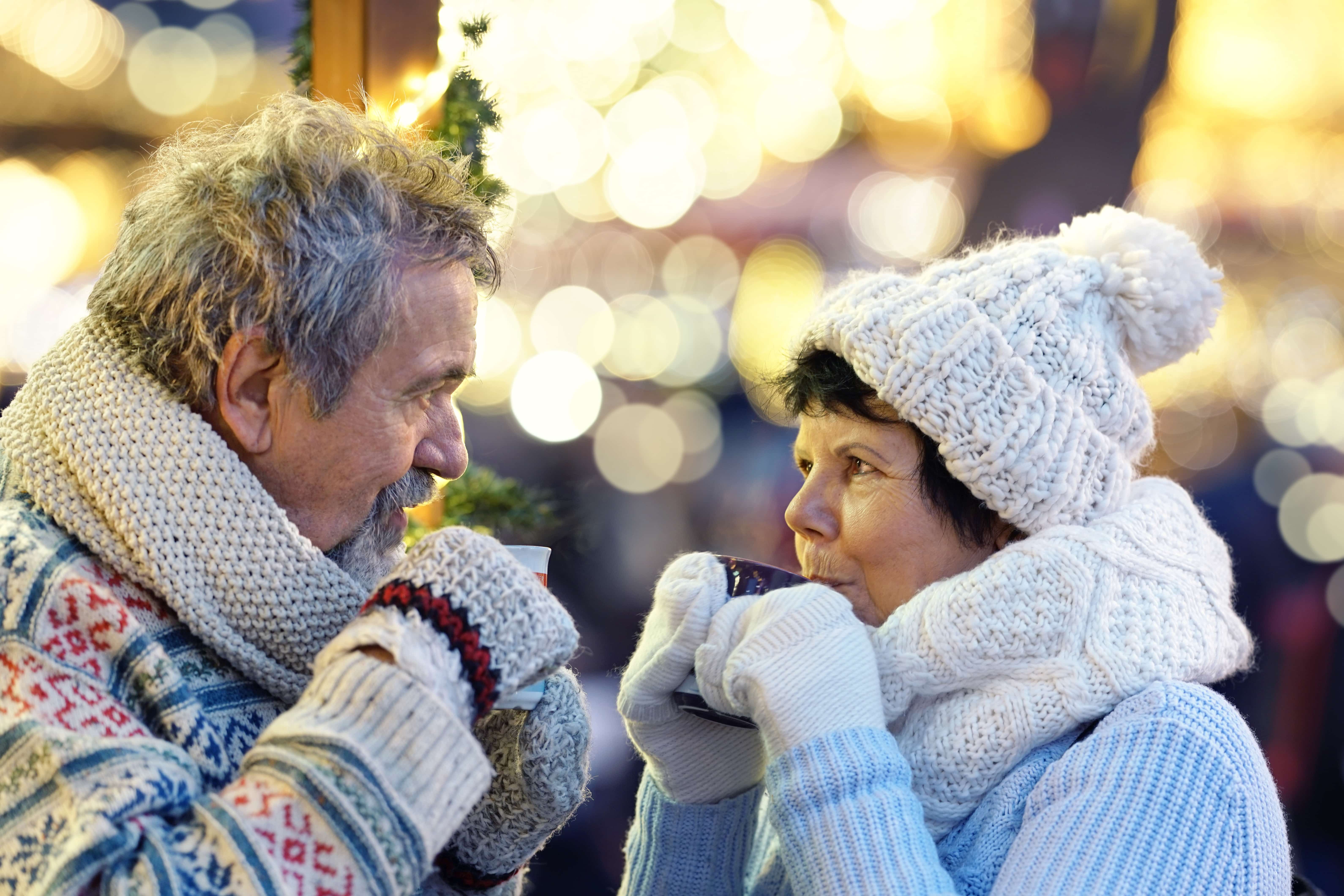 A senior man and woman wearing sweaters, gloves, hats, and scarves and drinking hot drinks to stay warm.