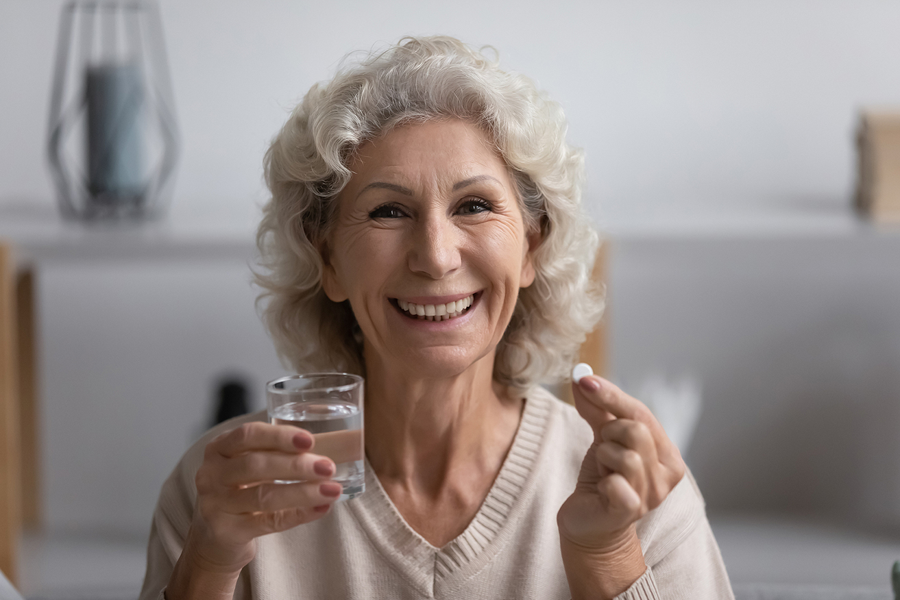 Senior woman smiling and holding a vitamin or medicine capsule and a glass of water up 