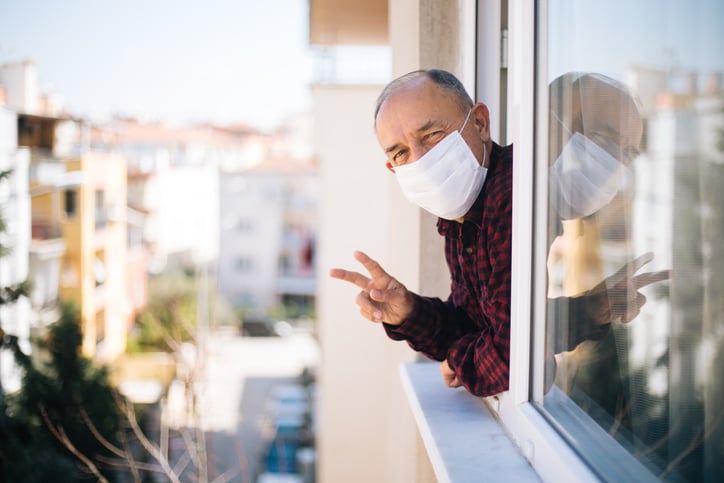 Senior man wearing face mask looking out window of senior living community home while sheltering in place