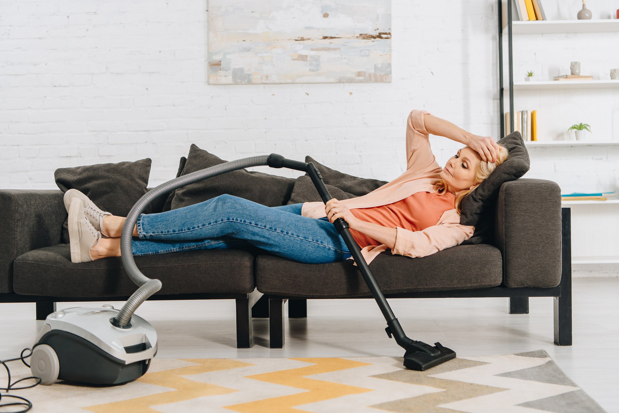Senior homeowner laying down on a couch holding the vacuum after cleaning the house and becoming exhausted